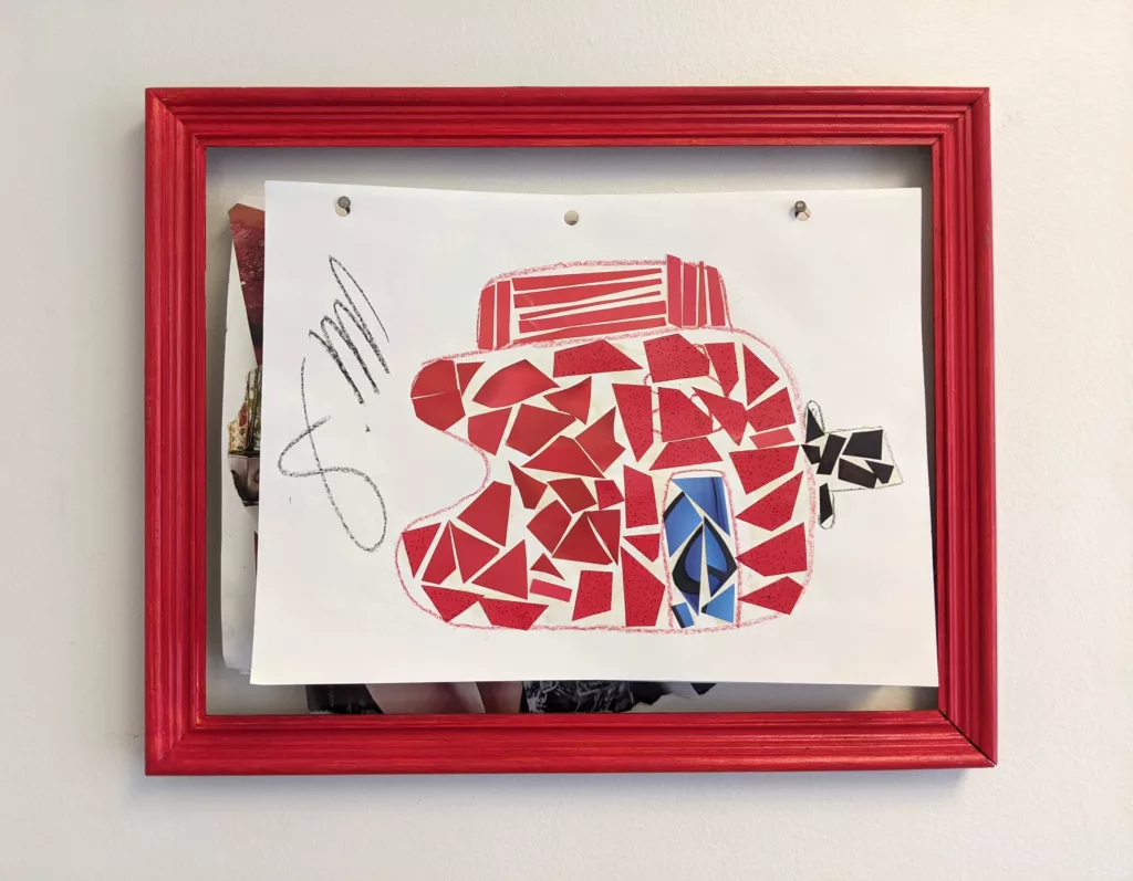 Red frame attached to wall with two nails just below the top of the frame. Paper artwork with hole punches are hung from the nails and displayed within the frame.