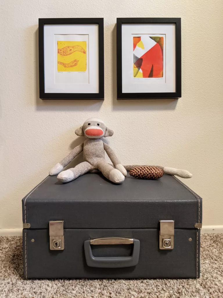 A sock monkey sits on a small suitcase with two small pieces of art hanging above it